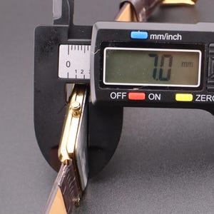 以下が含まれることがあります： 茶色の革バンドが付いた金色の腕時計をデジタルノギスで測定しています。ノギスは7.0mmの測定値を表示しています。この時計は長方形の文字盤とクラシックなデザインをしています。