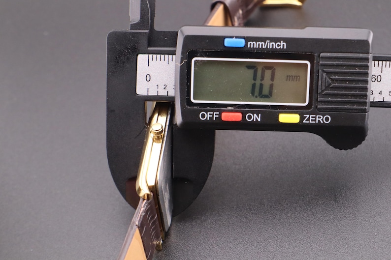 以下が含まれることがあります： 茶色の革バンドが付いた金色の腕時計をデジタルノギスで測定しています。ノギスは7.0mmの測定値を表示しています。この時計は長方形の文字盤とクラシックなデザインをしています。