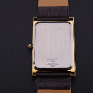 以下が含まれることがあります： 金色の長方形のセイコー製クォーツ腕時計。文字盤はシルバーで、茶色の革バンドが付いています。時計の裏側にはセイコーのロゴとその他の文字が見えます。時計は黒い面に置かれています。