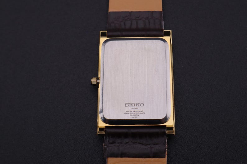 以下が含まれることがあります： 金色の長方形のセイコー製クォーツ腕時計。文字盤はシルバーで、茶色の革バンドが付いています。時計の裏側にはセイコーのロゴとその他の文字が見えます。時計は黒い面に置かれています。