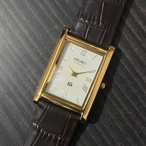 May include: A rectangular Seiko Quartz watch with a gold-tone case and a white dial featuring Roman numerals. The watch has a brown leather strap with a crocodile pattern. The watch face displays the Seiko Quartz logo.