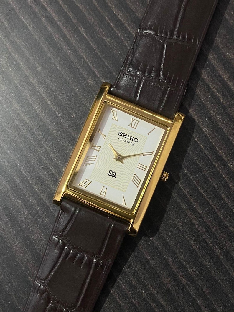 May include: A rectangular Seiko Quartz watch with a gold-tone case and a white dial featuring Roman numerals. The watch has a brown leather strap with a crocodile pattern. The watch face displays the Seiko Quartz logo.