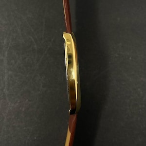May include: A gold-plated rectangular wristwatch with a brown leather strap. The watch face is visible, and the strap is attached to the watch case. The watch has a classic design.