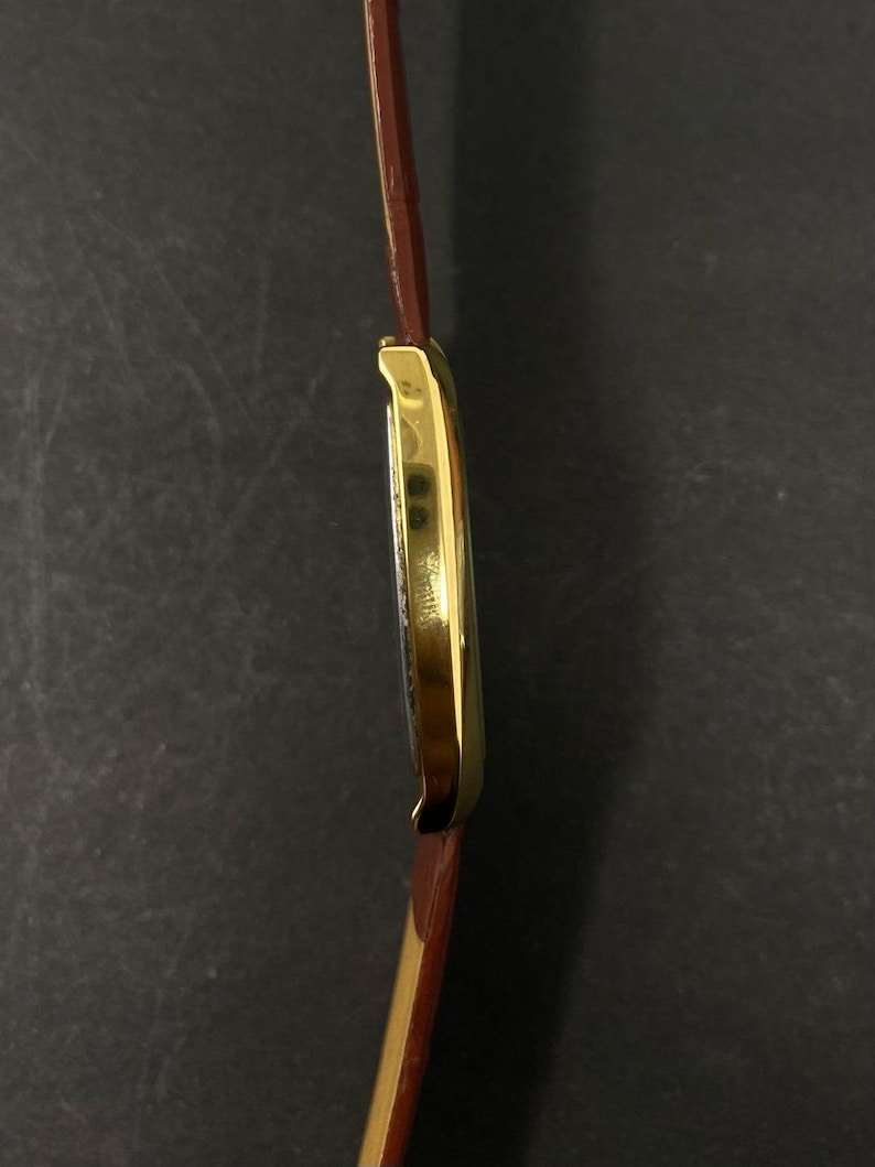 May include: A gold-plated rectangular wristwatch with a brown leather strap. The watch face is visible, and the strap is attached to the watch case. The watch has a classic design.