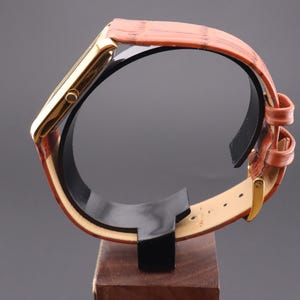 K&ouml;nnte beinhalten: Eine goldfarbene rechteckige Armbanduhr mit einem braunen Lederarmband. Die Uhr wird auf einem schwarzen St&auml;nder mit Holzsockel pr&auml;sentiert. Das Armband hat eine strukturierte Optik und eine goldene Schnalle.