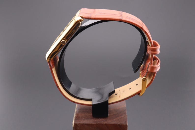 K&ouml;nnte beinhalten: Eine goldfarbene rechteckige Armbanduhr mit einem braunen Lederarmband. Die Uhr wird auf einem schwarzen St&auml;nder mit Holzsockel pr&auml;sentiert. Das Armband hat eine strukturierte Optik und eine goldene Schnalle.