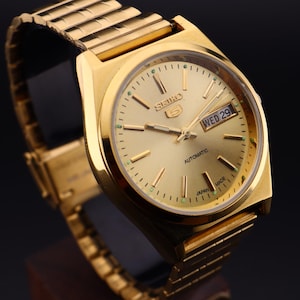 May include: A gold-tone Seiko 5 automatic wristwatch featuring a round gold dial with baton hour markers, luminous hands, and a day-date window at the three o'clock position. The watch includes a matching gold-tone metal link bracelet strap.