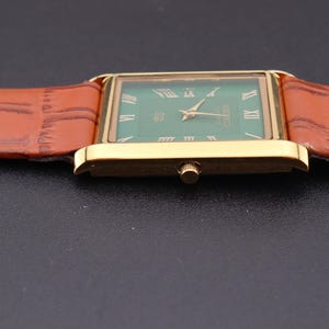 K&ouml;nnte beinhalten: Eine rechteckige Armbanduhr in Gold mit gr&uuml;nem Zifferblatt und r&ouml;mischen Ziffern. Die Uhr hat goldene Zeiger und ein braunes Lederarmband. Das Armband hat ein strukturiertes, krokodilartiges Muster.