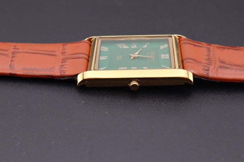 K&ouml;nnte beinhalten: Eine rechteckige Armbanduhr in Gold mit gr&uuml;nem Zifferblatt und r&ouml;mischen Ziffern. Die Uhr hat goldene Zeiger und ein braunes Lederarmband. Das Armband hat ein strukturiertes, krokodilartiges Muster.
