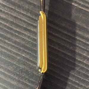 May include: A gold-toned rectangular wristwatch with a brown leather strap. The watch case has a sleek, minimalist design. The watch is seen from the side, showcasing its thin profile and the contrast between the gold and the dark strap.
