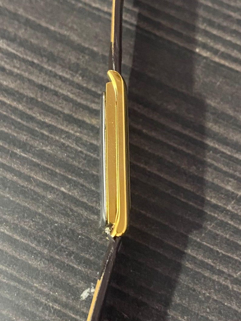 May include: A gold-toned rectangular wristwatch with a brown leather strap. The watch case has a sleek, minimalist design. The watch is seen from the side, showcasing its thin profile and the contrast between the gold and the dark strap.