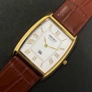 May include: A gold-plated Seiko Quartz watch with a rectangular white face, Roman numerals, and gold hands. The watch has a brown leather strap with a crocodile pattern. The watch face displays the Seiko logo.