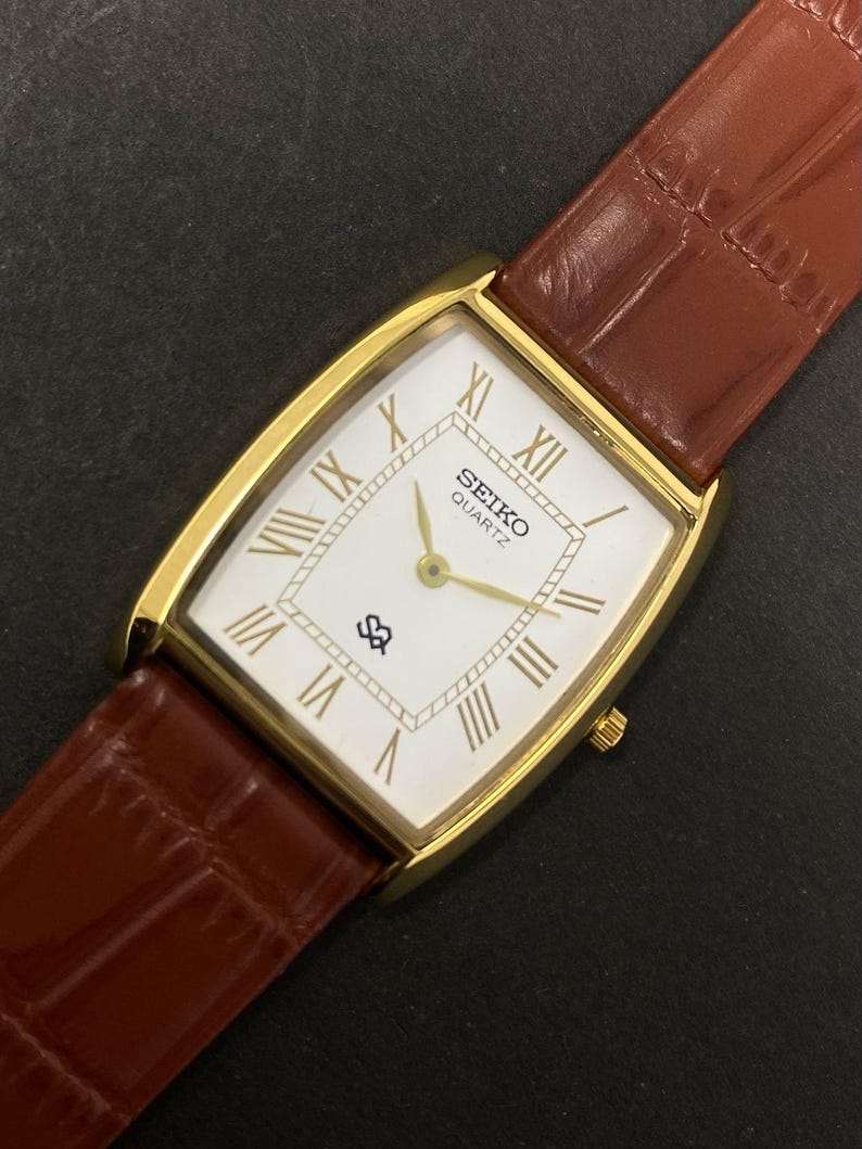 May include: A gold-plated Seiko Quartz watch with a rectangular white face, Roman numerals, and gold hands. The watch has a brown leather strap with a crocodile pattern. The watch face displays the Seiko logo.