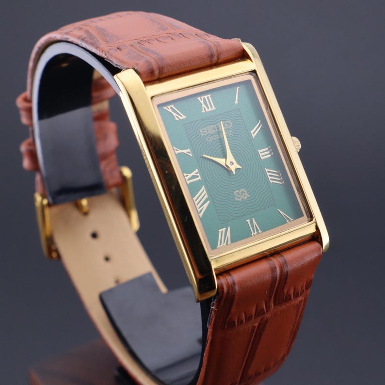 K&ouml;nnte beinhalten: Eine rechteckige, goldfarbene Armbanduhr mit gr&uuml;nem Zifferblatt und r&ouml;mischen Ziffern. Das Armband ist aus braunem Leder mit Krokodilmuster. Auf dem Zifferblatt sind das Seiko-Logo und die Buchstaben "sa" zu sehen.