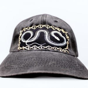 May include: A black baseball cap with a white snake skeleton embroidered on a black patch. The patch is surrounded by a brown stitched border.