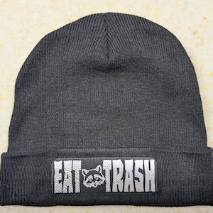 May include: A dark gray beanie hat with a rectangular patch that reads "EAT TRASH" in silver lettering. The patch features a raccoon graphic. The hat is made of a ribbed knit material.