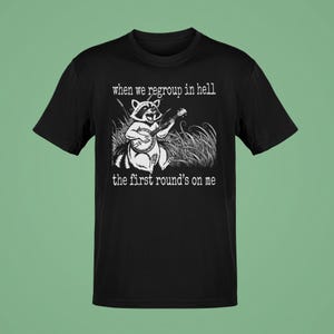 May include: Black t-shirt with a white graphic of a raccoon playing a banjo in tall grass. The text reads "when we regroup in hell" above the raccoon and "the first round's on me" below.