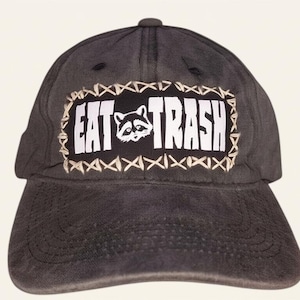May include: A dark gray baseball cap with a curved brim. The front features the text "EAT TRASH" in white block letters, with a raccoon graphic. The text is framed by a decorative stitched border.