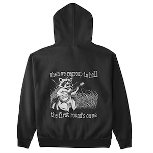 May include: Black hooded sweatshirt with a white graphic of a raccoon playing a banjo, surrounded by tall grass. Text above reads "when we regroup in hell" and below "the first round's on me."