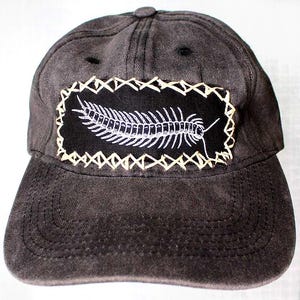 May include: A black baseball cap with a white embroidered centipede design on a black patch. The patch is surrounded by a beige stitched border.