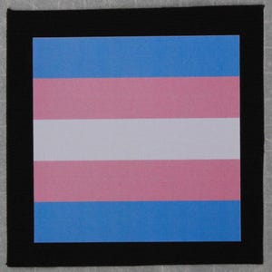 May include: A square patch featuring the transgender pride flag, with horizontal stripes of light blue, pink, and white. The flag is framed by a black border, creating a bold contrast. The design is simple and impactful.