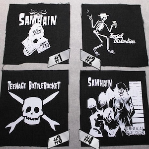 May include: Four black and white iron-on patches. The first patch features the band name "SAMHAIN" in white text. The second patch features a skeleton in a hat with the text "Social Distortion". The third patch features a skull and crossbones with the text "TEENAGE BOTTLEROCKET". The fourth patch features a band of figures with the text "SAMHAIN" and "UNHOLY PASSION".