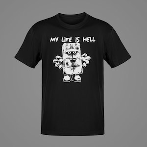 May include: Black t-shirt with a white graphic of a blocky, cartoonish figure with the text "MY LIFE IS HELL" above it. The figure has a rectangular body, round eyes, and stylized arms and feet.