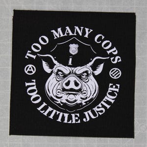 May include: Black and white patch with a pig wearing a police hat. The text around the pig says "Too Many Cops Too Little Justice".
