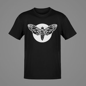 May include: Black t-shirt featuring a white graphic of a moth with a skull-like design on its body, set against a white circular background. The moth's wings are detailed with intricate patterns. The shirt has short sleeves and a crew neck.