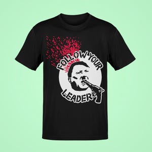 May include: Black t-shirt with a graphic design. The design features a black and white illustration of a person with a gun in their mouth, surrounded by the words "FOLLOW YOUR LEADER" in a white circle, with a red splatter effect.