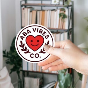 May include: A round sticker with the text "ABA VIBES CO." encircling a red heart with a smiling face. The sticker has a white background and a dark brown border with leaf accents. The sticker is held in a hand.