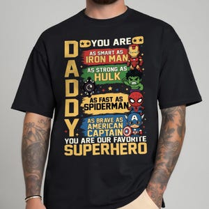 May include: Black t-shirt with a colorful graphic featuring the word "DADDY" and the phrase "YOU ARE OUR FAVORITE SUPERHERO." The design includes illustrations of Iron Man, Hulk, Spiderman, and Captain America.
