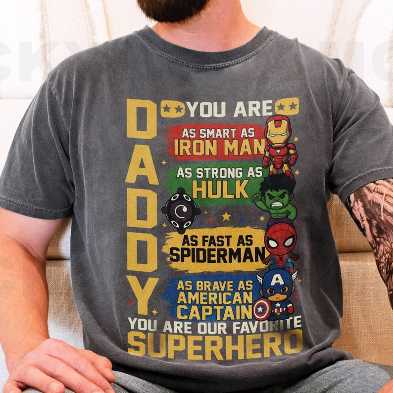 May include: Dark gray t-shirt with the word "DADDY" in yellow block letters. The shirt features illustrations of Iron Man, Hulk, Spiderman, and Captain America with the text "YOU ARE OUR FAVORITE SUPERHERO."