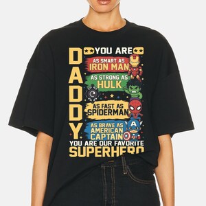 May include: Black t-shirt with the text "DADDY YOU ARE" and superhero references. The shirt features illustrations of Iron Man, Hulk, Spiderman, and Captain America, with text describing each superhero's qualities. The phrase "YOU ARE OUR FAVORITE SUPERHERO" is also included.