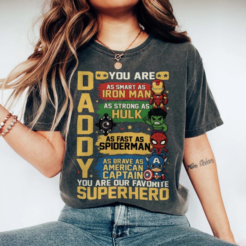 May include: Dark gray t-shirt with the text "DADDY" and "YOU ARE OUR FAVORITE SUPERHERO." The shirt features colorful illustrations of Iron Man, Hulk, Spiderman, and Captain America. A great gift for Father's Day.
