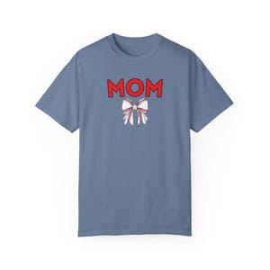 Peut inclure: Un t-shirt bleu clair avec le mot "MOM" en lettres rouges et un n&oelig;ud blanc avec des coutures rouges, ressemblant &agrave; un ballon de baseball, au centre.