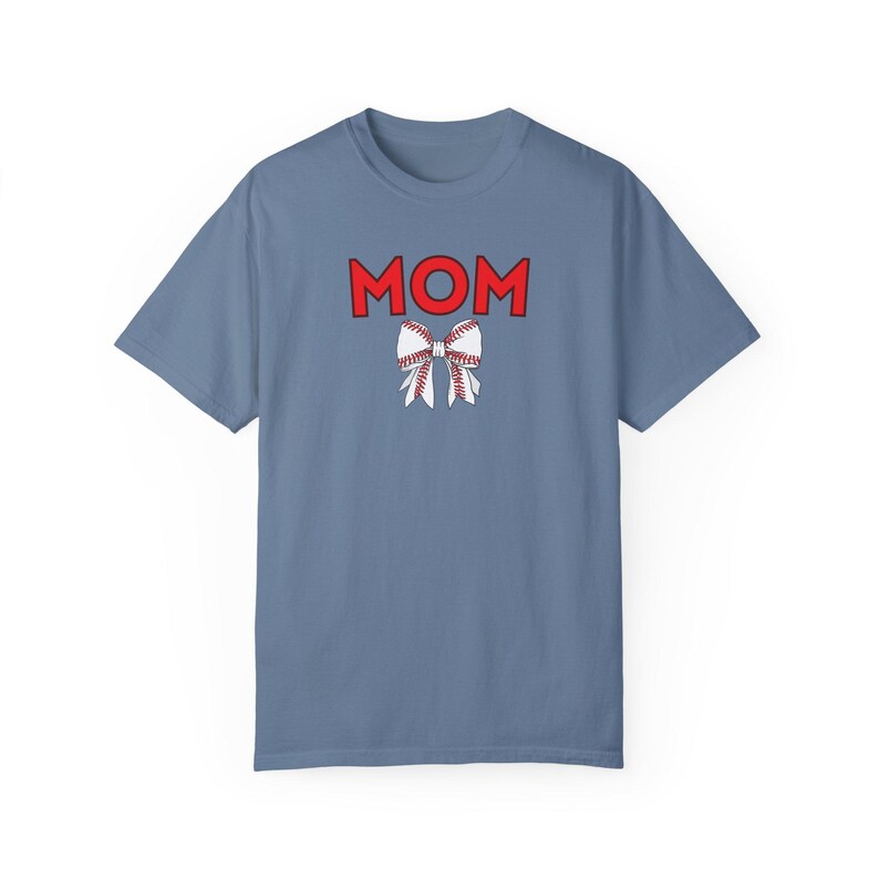 Peut inclure: Un t-shirt bleu clair avec le mot "MOM" en lettres rouges et un n&oelig;ud blanc avec des coutures rouges, ressemblant &agrave; un ballon de baseball, au centre.