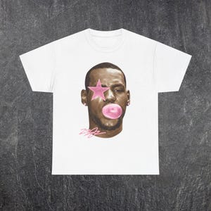 Puede incluir: Camiseta blanca con un retrato de una persona con una estrella rosa sobre un ojo y una burbuja de chicle. El retrato está en tonos marrones y rosas, con la firma "Zzz" en rosa.