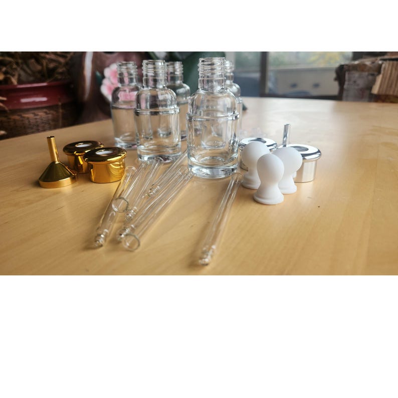 Puede incluir: Un juego de botellas de vidrio transparente con tapas doradas y goteros, junto con dos embudos dorados y dos tapones de goma blancos. Las botellas est&aacute;n dispuestas sobre una superficie de madera clara.