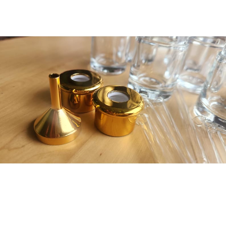 Puede incluir: Tapones de botella dorados con un embudo. Los tapones tienen un anillo interior blanco y un anillo exterior dorado. El embudo es dorado y tiene un pico estrecho.