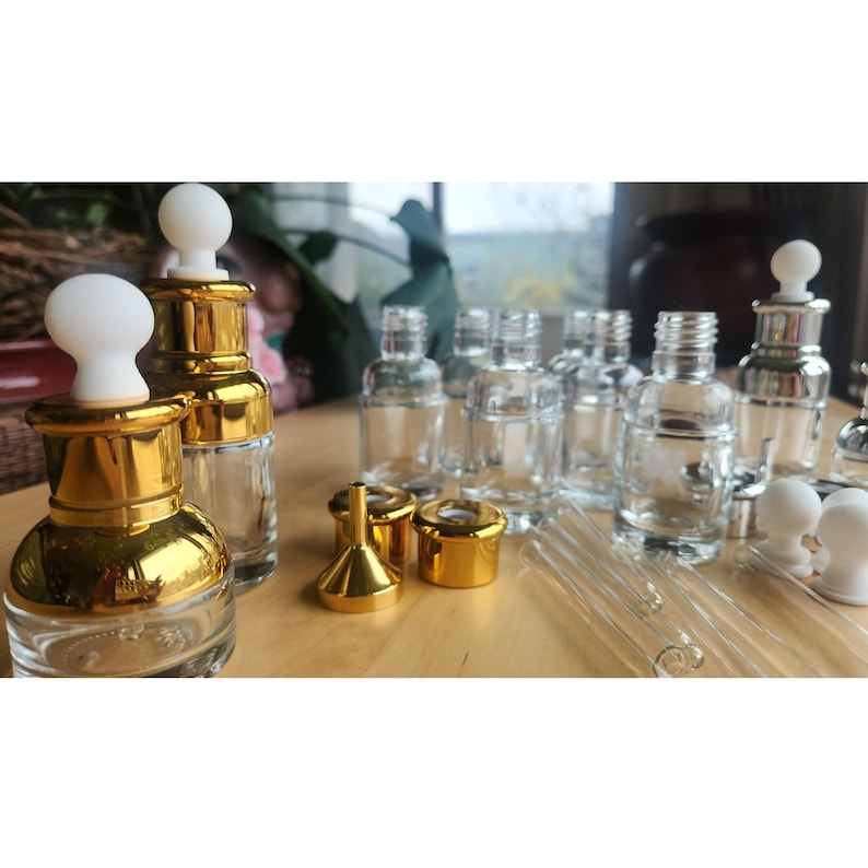 Puede incluir: Un juego de botellas de vidrio transparente con tapas doradas y goteros, junto con dos embudos dorados. Las botellas est&aacute;n dispuestas sobre una superficie de madera.