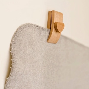 May include: A wooden blanket holder mounted on a wall, featuring a heart-shaped accent. The holder is made of light-colored wood and is holding a gray blanket. The blanket has a stitched edge.