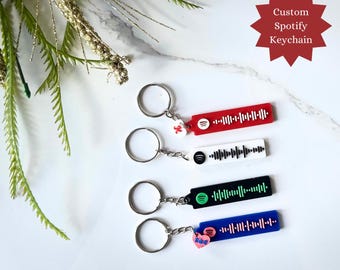 Pode incluir: Quatro pingentes personalizados do Spotify com fundos de cores diferentes. Os pingentes são vermelho, branco, preto e azul. Cada pingente tem um código do Spotify. O texto "Custom Spotify Keychain" está em forma de estrela acima dos pingentes.