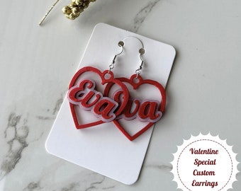 Op de afbeelding: Rode hartvormige oorbellen met de naam "Eva" in wit erin geschreven. De oorbellen bevinden zich op een witte kaart met de tekst "Valentine Special Custom Earrings".