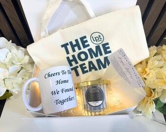Pode incluir: Um conjunto de presentes com uma caneca branca com o texto "Cheers To The Home We Found Together", uma vela num frasco de vidro, uma sacola com "THE HOME TEAM" impresso e um cartão. O conjunto é apresentado numa caixa branca.
