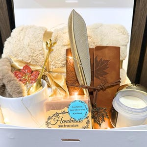 May include: A gift basket with a variety of items, including a plush blanket, a mug with a decorative spoon, a candle, a journal, and a jar of cream. The basket also contains a decorative feather and a bar of soap. The soap label reads "Handmade come from nature."