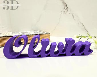 Könnte beinhalten: Ein violettes 3D-Namensschild mit der Aufschrift "Olivia". Das Schild besteht aus einem glatten, festen Material und hat eine leicht gekrümmte Form.