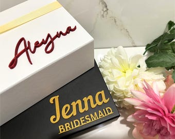 May include: Two gift boxes with personalized names. The top box is white with a red cursive name "Aleyna". The bottom box is black with a gold cursive name "Jenna" and the text "BRIDESMAID".