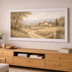 Puede incluir: Un televisor grande con marco blanco muestra una pintura de paisaje de una escena de la campiña toscana. El televisor está montado sobre un mueble de madera clara con varios cajones y estantes. También se ve un jarrón con flores blancas, velas y libros.