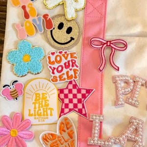 May include: A white canvas tote bag with pink straps, decorated with various colorful patches including a glittery smiley face, a pink checkered star, a blue flower, a bee, and text patches reading God Is Love, Love Your Self, and Be The Light.